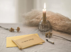 Afbeelding van een wenskaartvormige kaars die op tafel ligt waar de woorden 'ik denk aan je' op geschreven zijn. Er naast ligt een krijtje, gele droogbloemen en een glazen kandelaar waar een brandende Wenskaars in zit.